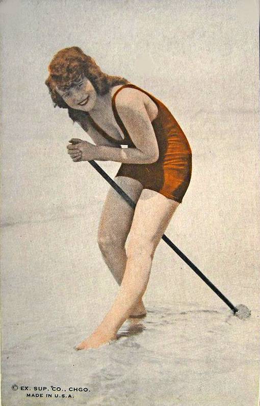 ARCADE CARD - EXHIBIT SUPPLY COMPANY - RED-HAIRED WOMAN STANDING IN SHALLOW WATER WITH A TOOL WEARING RED WOOLLY BATHING SUIT - TINTED - 1920s
